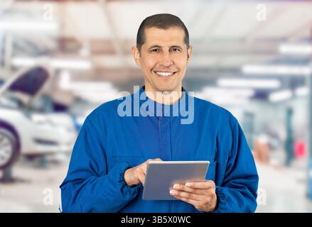 Shot of confident smiling professional handyman in blue overalls using digital tablet at repair shop. Stock Photo