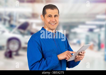 Confident smiling professional handyman in blue overalls using digital tablet at repair shop. Stock Photo