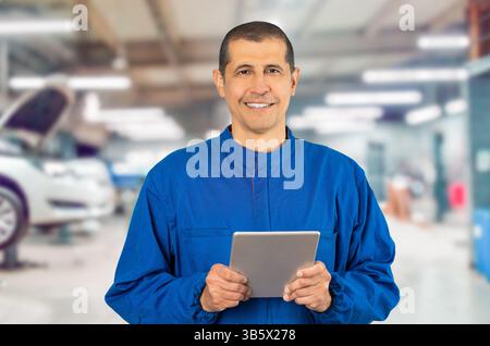 Confident smiling professional handyman in blue overalls using digital tablet and looking a camera at repair shop. Stock Photo