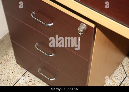 A close-up view of a wooden desk with three drawers. The drawers have three metallic handles and the top one has security lock with keys. Stock Photo