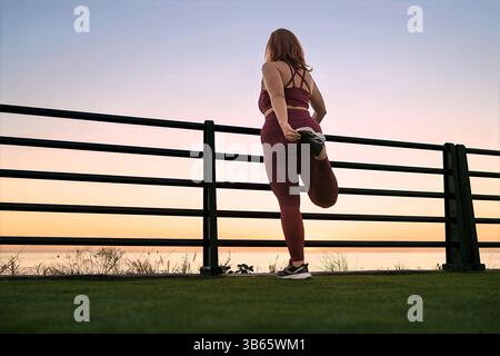 Active woman doing leg stretch at sunrise by the sea. Healthy outdoor fitness routine and wellness concept. Stock Photo