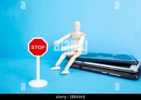 Wooden mannequin sitting on a smartphones stack with a stop sign on blue background Stock Photo