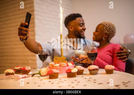 Happy young couple celebrating woman's birthday at home. They are embracing and taking selfie. Selective focus on cake. Stock Photo