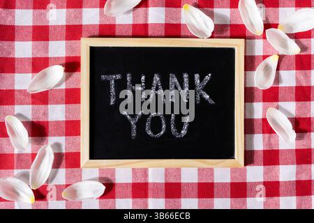 A small chalkboard displaying a Thank You message surrounded by delicate white petals, placed on a red and white checkered tablecloth for expressing g Stock Photo
