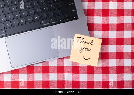 Simple remote workspace featuring a laptop and a bright yellow Thank you note on a red and white checkered tablecloth, perfect for conveying gratitude Stock Photo