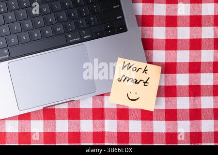 Sticky note with "Work smart" message and smiley face placed next to a laptop on a red and white checkered surface, symbolizing simple motivation for Stock Photo