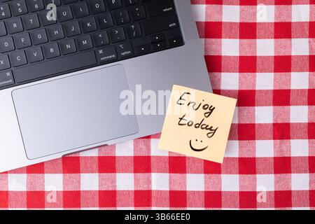 Sticky note with "Enjoy today" text and smiley face placed next to a laptop on a red and white checkered surface, symbolizing a positive reminder for Stock Photo