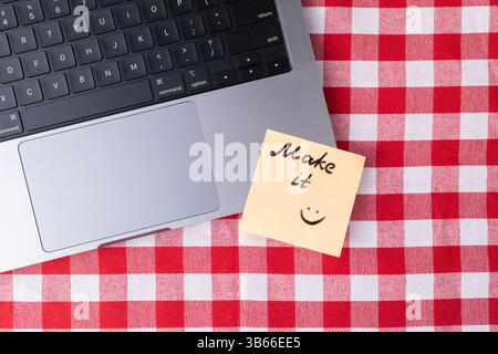 Yellow sticky note with handwritten Make it message and smiling face is placed near a laptop on a red checkered tablecloth representing motivation, pr Stock Photo