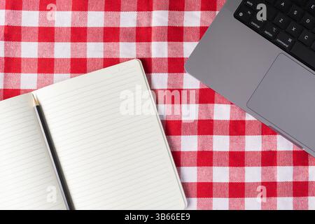Open blank notebook, pencil, and laptop keyboard on a red checkered tablecloth surface, symbolizing remote work, home office, planning, digital workfl Stock Photo