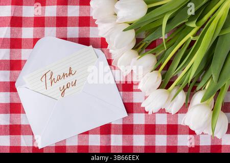 Thank you message on a card in an envelope arranged with white tulips on a red checkered tablecloth background, ideal for conveying gratitude and appr Stock Photo