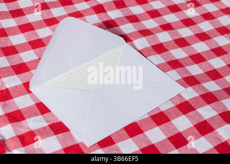 Open white envelope with blank lined card inside, on red checkered tablecloth background, perfect for adding messages, invitations, or greetings Stock Photo