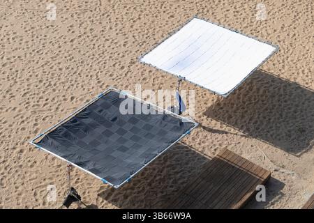 Two large reflectors are set up on a sandy beach. One is white and the other is black. control lighting during outdoor photo or film shoots. Stock Photo