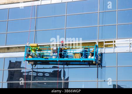 Workers are utilizing a lift to safely clean the windows of a tall glass skyscraper in an urban environment Stock Photo