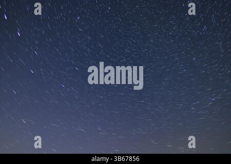 Beautiful startrails at dark night sky over some fields and high trees silhouettes at sky with milky way with moving clouds and flying Perseids Stock Photo