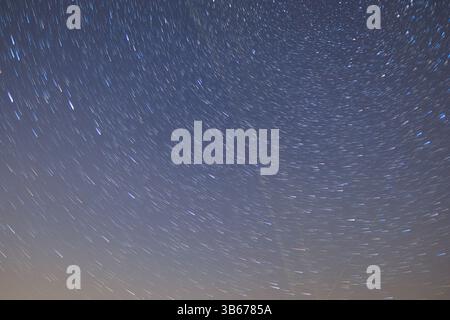 Beautiful startrails at dark night sky over some fields and high trees silhouettes at sky with milky way with moving clouds and flying Perseids Stock Photo