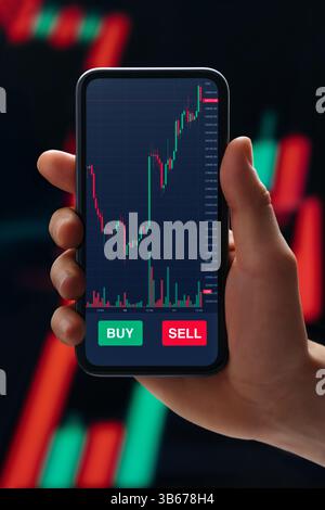 Close up, trader hand holding smartphone with stock market app on screen, against candlestick diagram on background. Online trading and investment concept Stock Photo