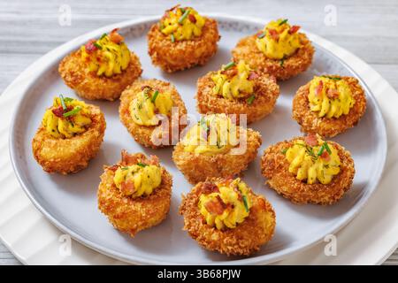 Deep fried deviled eggs with crispy coating of panko and parmesan, sprinkled with fresh chives and crumbled bacon on white plate on wooden table, clos Stock Photo