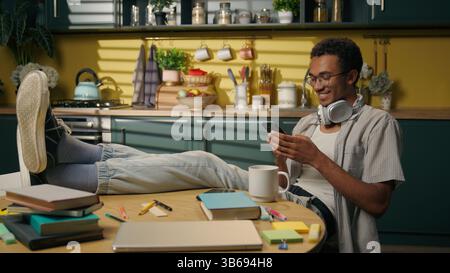 African American smiling guy ethnic male freelancer student relax studying break playing online game smartphone usage at home kitchen relaxed gen z Stock Photo