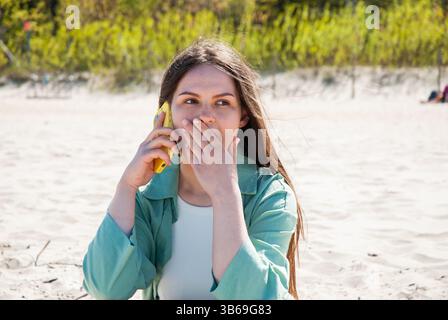 Young caucasian woman at the beach in a green jacket covers her mouth while talking on a yellow phone, expressing surprise and contemplation. Digital Stock Photo