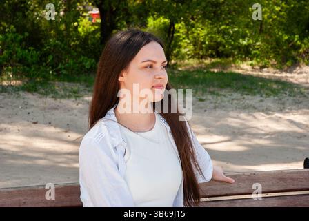Young caucasian female in a white shirt with long brown hair sits pensively on a park bench surrounded by greenery and sand.. Digital nomad lifestyle. Stock Photo