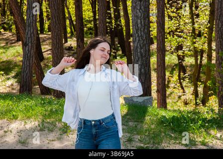 Young caucasian woman enjoying music outdoors in a serene forest setting, wearing casual attire with a relaxed expression and earphones. Digital nomad Stock Photo