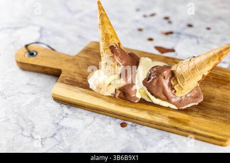 Horizontal image of two ice-cream cones placed upside down on a wooden board. The ice cream forms creamy puddles that contrast with the light marble s Stock Photo