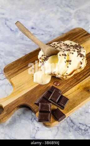 Vertical image of a cup of partially melted vanilla ice cream, with chocolate shavings and a chopped chocolate bar next to it. All on a rustic wooden Stock Photo