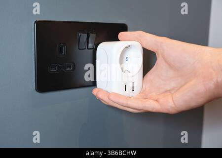 Hand holding a smart power EU adapter with USB ports next to a black UK outlet Stock Photo