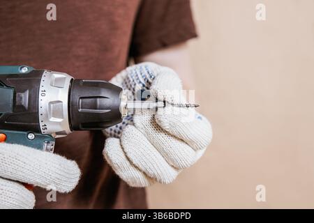 Gloved hands hold a power drill and metal screw in close-up against neutral background with copy space Stock Photo