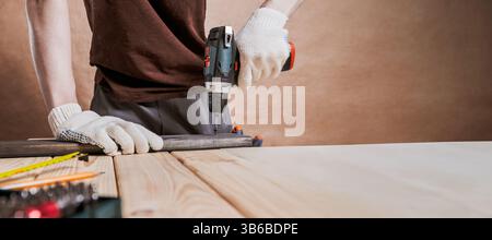 Man in gloves using electric drill on wood plank over table in workshop. DIY ,repair, home improvement Stock Photo
