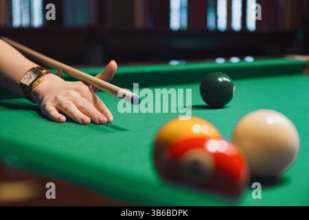 Close-up of hand holding cue stick aiming to strike billiard balls on a green felt pool table. Sports, leisure activity Stock Photo