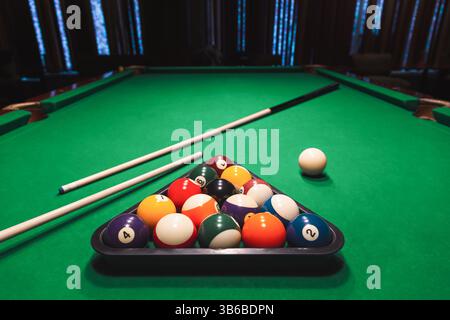 Full set of numbered pool balls arranged in a triangle rack with cue sticks on a green pool table. Bar games, cue sports Stock Photo