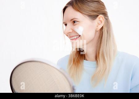 Young woman with blonde hair smiling with white heart shape cream on cheek, applying moisturize cream, beauty routine Stock Photo