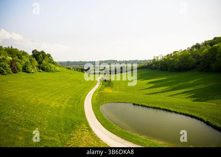Aerial perspective of a winding country path meandering through vibrant green rolling hills and fields, flanked by trees under a clear sky. Stock Photo