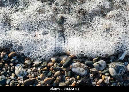 Soft white sea foam is covering small colorful pebbles on a beach Stock Photo