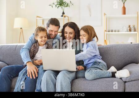 Smiling parents sitting on sofa with kids, using laptop and enjoying fun time together at home. Stock Photo