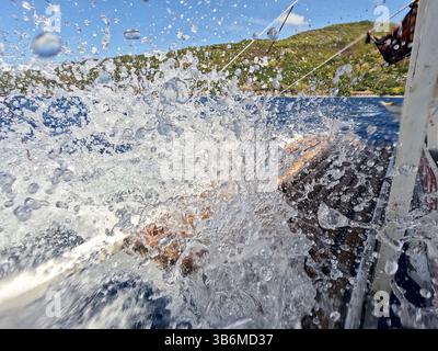 Dramatic splash of seawater captured from the side of a moving boat with scenic green hills and blue ocean in the background, evoking adventure. Stock Photo