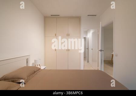 Minimalist bedroom with taupe bedding, white built-in wardrobe and two open doorways Stock Photo