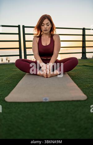 Serene young woman meditating on yoga mat at seaside park during sunset, relaxing and practicing mindfulness outdoors Stock Photo