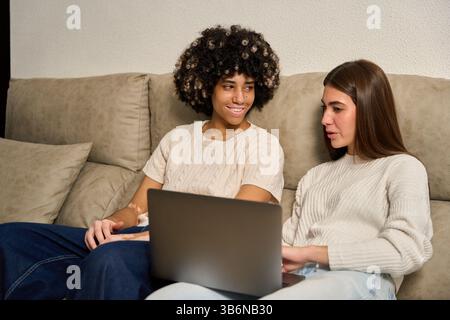 Two female students collaborating on laptop, studying together on sofa Stock Photo