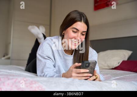 Happy female student using smartphone while lying on bed Stock Photo