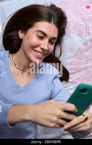 Happy female student using smartphone lying on bed Stock Photo