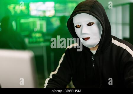 Hacker working on laptop in dark room with green code displayed on monitors Stock Photo