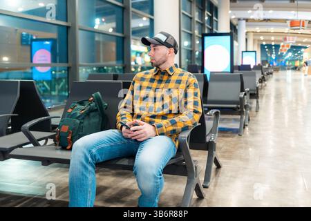 Mid shot of adult Caucasian man in cap and jacket using smartphone in airport waiting area sitting in chair. Concept of boarding at terminal before fl Stock Photo