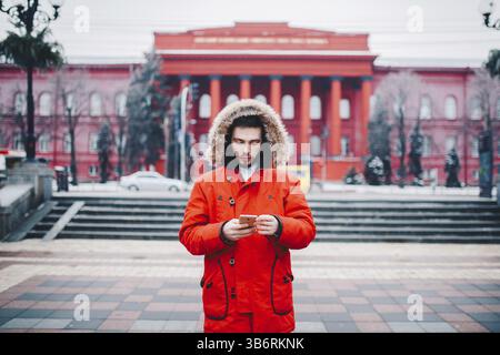 A handsome young guy with beard and red jacket in hood a student uses a mobile phone, writes, writes a correspondence over the phone with a smile agai Stock Photo