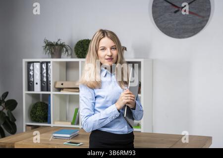 Businesswoman Holding Large File. young womanl standing in the office and holding folder. portrait of young office worker holding folders of paperwork Stock Photo
