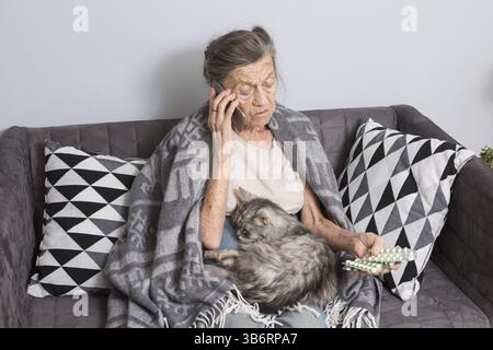 Sick senior woman taking medicine at home. Senior woman talking on mobile phone while looking at medicine in bedroom. pensioner studying medicine call Stock Photo
