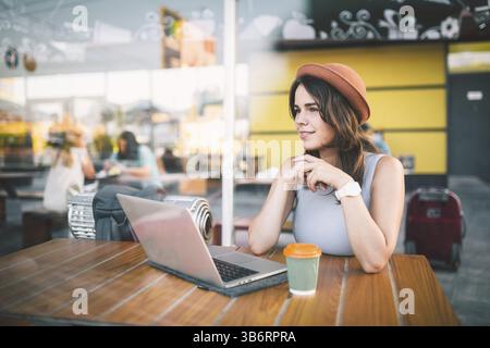 Beautiful young caucasian woman sitting on a terrace in a cafe in summer at a wooden table in a hat and a plate uses technology, working behind a lapt Stock Photo