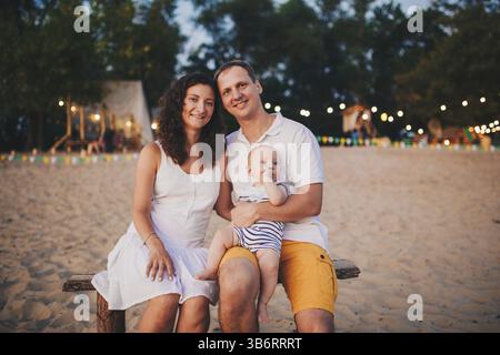 Theme of family summer holidays on the beach. Caucasian man and woman dad and mom hug a small baby son sitting on his hands. Young family relaxing in Stock Photo