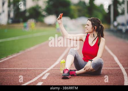 Young woman athlete at the stadium sporting lifestyle, sitting on the track, taking selfie photo on a smartphone, smiling joyful in her ears earpiece Stock Photo